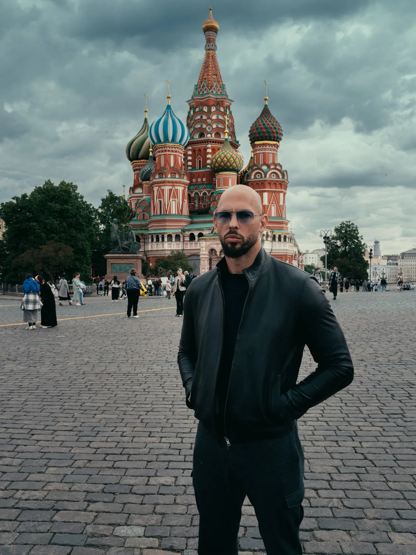 Andrew Tate in a black leather jacket and sunglasses standing in Red Square in front of Saint Basil's Cathedral in Moscow